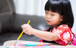Child holding brush in her left hand is painting water color on paper