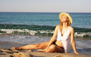Girl in straw hat and white swim suit sitting on the beach sunbathing